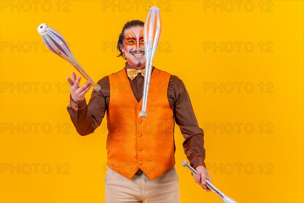 Happy juggler man in makeup vest juggling maces on a yellow background