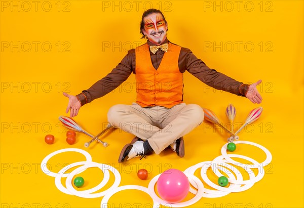 Portrait of a juggler in a vest and makeup sitting with the juggling objects on a yellow background