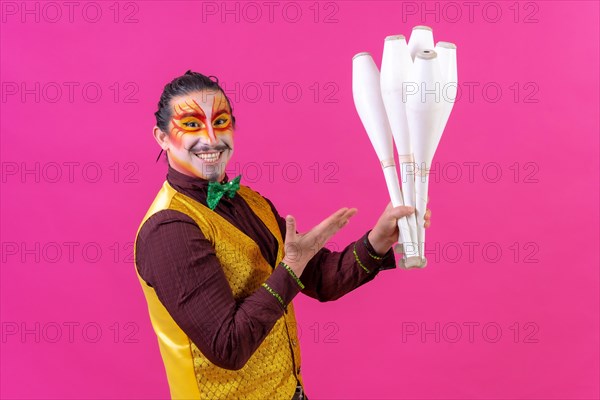 Juggler in a vest and with painted face juggling maces on a pink background