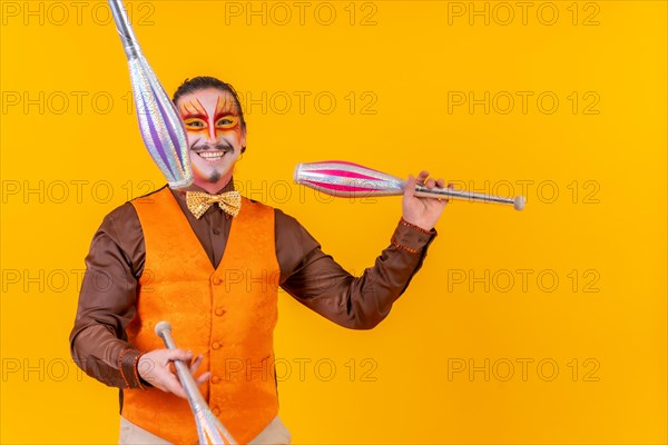 Juggler man in make up vest juggling with maces on a yellow background