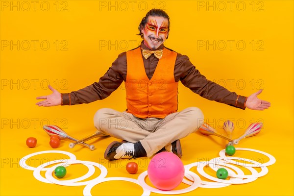 Juggler in a vest and with his face painted sitting with the juggling objects on a yellow background