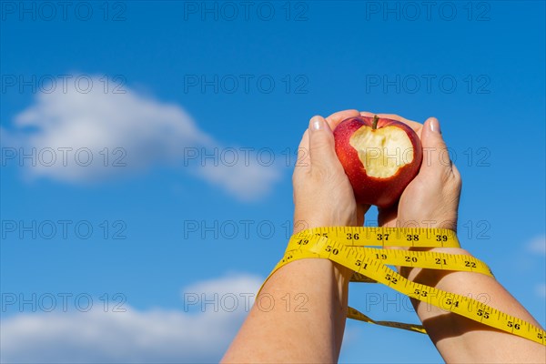 Woman holding a bitten apple in her hands with a tape measure and a sky with clouds in the background