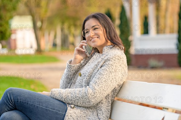 Pretty woman with glasses and Iphone in hand sits on bench in park