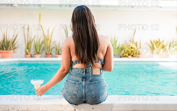 Woman on vacation sitting on the edge of swimming pool with a drink