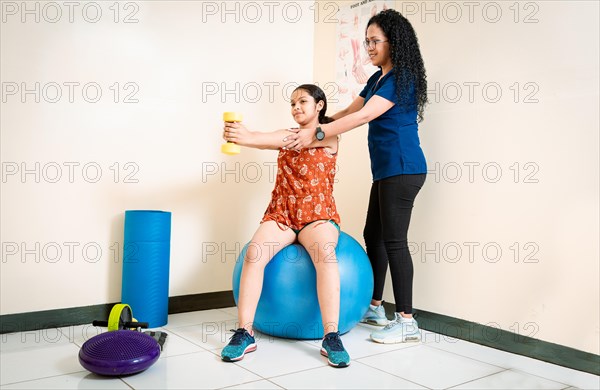 Physiotherapist helping patient woman with dumbbell sitting on rehabilitation ball. Chiropractor helping woman patient with dumbbell sitting on Fitness Ball