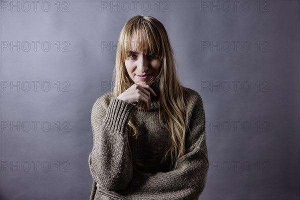 Woman with long blond hair and woollen jumper holds a hand to her chin and smiles at camera