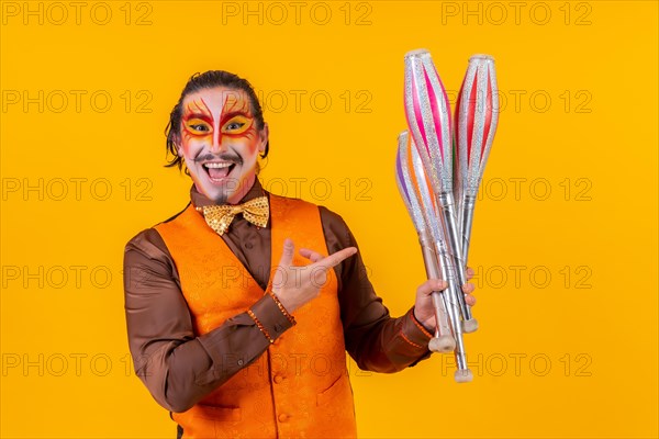 Portrait of a happy juggler man in make up vest juggling with maces on a yellow background
