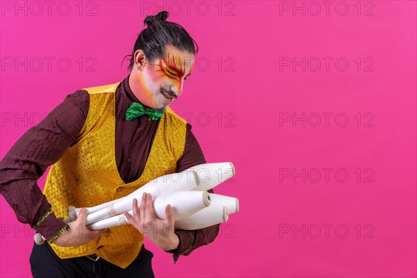 Juggler in a vest and with painted face juggling clubs on a pink background