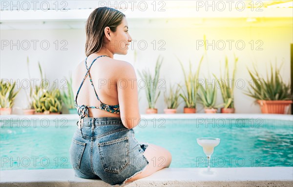 Girl sitting on the edge of the pool with tropical drink. Rear view of girl sitting on the edge of the pool with a drink. Woman on vacation sitting on the edge of swimming pool with a drink