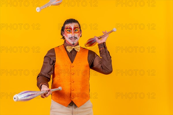 Happy juggler man in makeup vest juggling maces on a yellow background