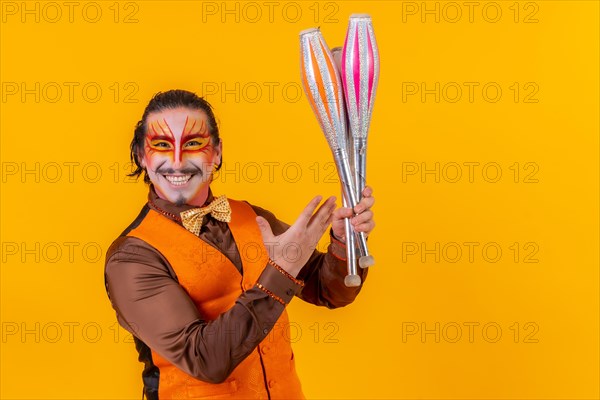 Portrait of a juggler in a vest with a painted face juggling with maces on a yellow background