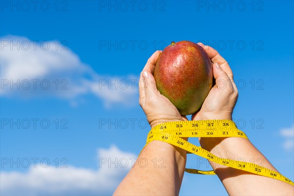 Woman holding a mango in her hands with a tape measure and a sky with clouds in the background