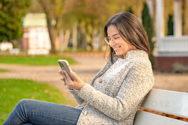 Pretty woman with glasses and Iphone in hand sits on bench in park