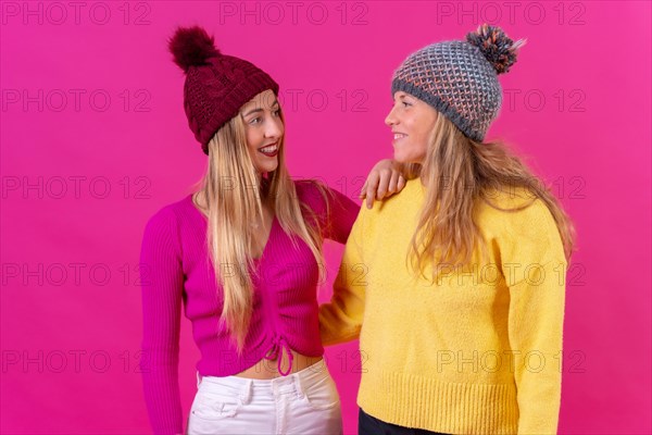 Two blonde caucasian women in wool hats on a pink background looking at each other smiling