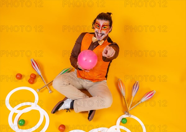 Portrait of a juggler in a vest and makeup sitting with the juggling objects on a yellow background