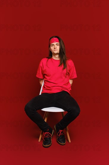 Young adult man in black and red casual clothes sits on chair with his arms behind back