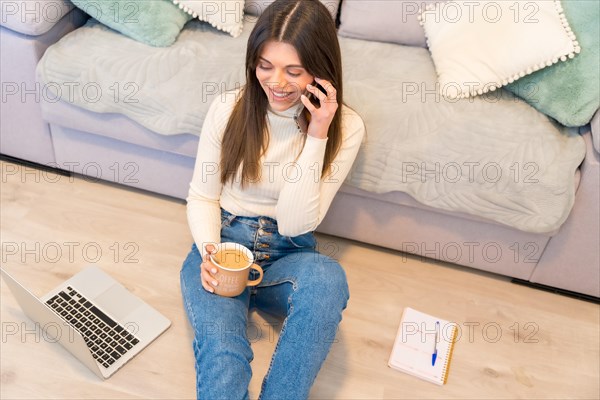 Portrait of woman with a computer sitting on a sofa with a hot coffee talking on the phone