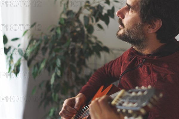 Young man with beard playing Spanish guitar looking out the window with sunlit face