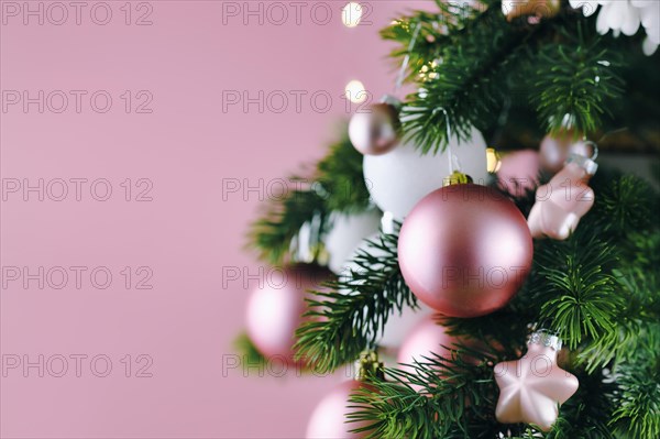 Close up of decorated Christmas tree with white seasonal and pink tree ornaments like baubles and stars on pink background with lights in background
