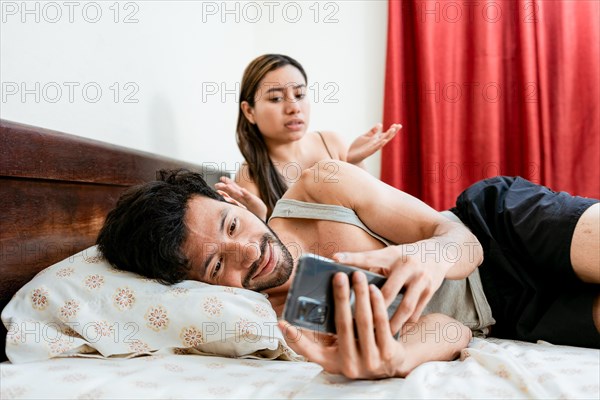 Husband with cell phone ignoring wife in bed. Annoyed woman with husband while holding cell phone in bed. Cheating Husband Texting On Phone Ignoring Wife Lying In Bed