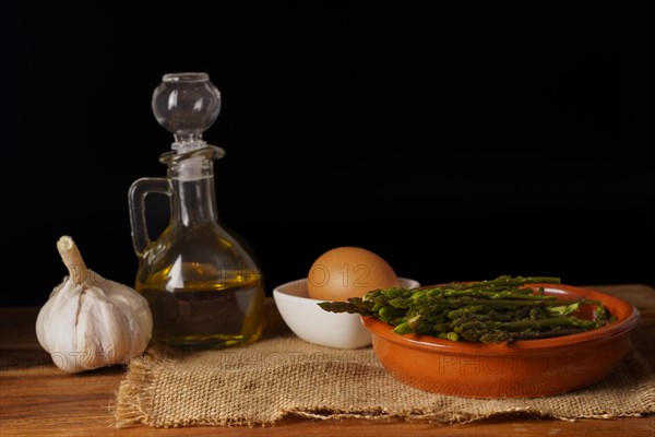 Bunch of wild asparagus on a raffia cloth with a jar of olive oil and eggs black background