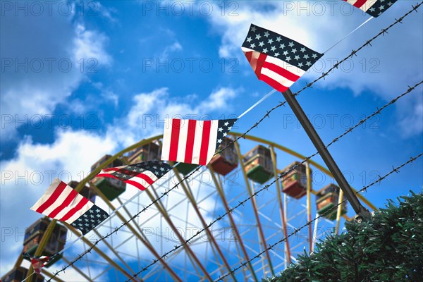 Simplified flag with American colors with red stripes and white stars on blue background hanging next to a barbed wire fence with ferris wheel in background