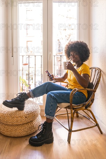 Afro hair woman by the window home having a hot drink, coffee in the morning looking at the mobile