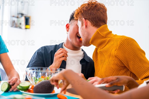 Group of friends preparing vegetarian food. Preparing the salad and having fun in the kitchen, lgtb boy couple kiss with carrot