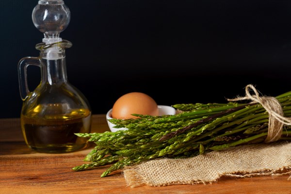 Bunch of wild asparagus on a raffia cloth with a jar of olive oil and eggs black background