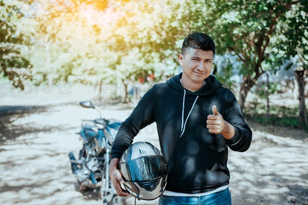Portrait of motorcyclist holding safety helmet with thumb up. Smiling motorcyclist man holding safety helmet on the road. Motorcyclist giving thumbs up holding safety helmet on the road