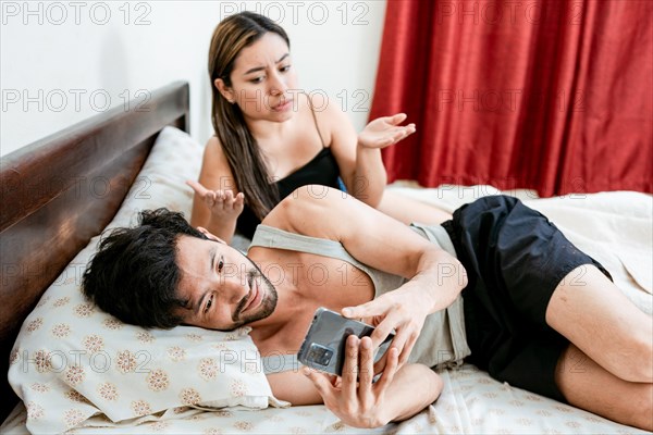 Cheating Husband Texting On Phone Ignoring Wife Lying In Bed. Husband with cell phone ignoring wife in bed. Annoyed woman with husband while holding cell phone in bed