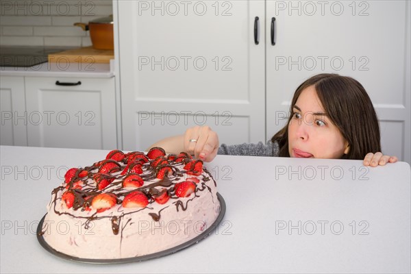 Young woman hiding behind the table eating strawberry shortcake in her home kitchen