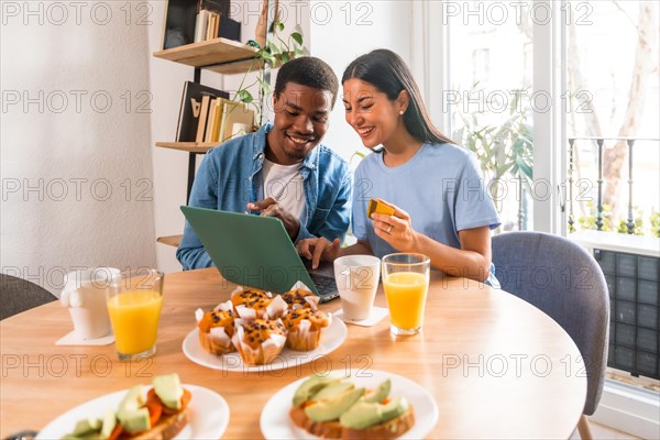 Couple making purchase online with the computer while having breakfast, next to the window, booking the holidays