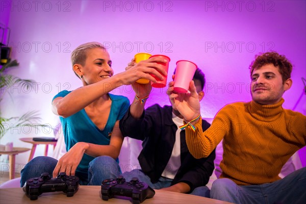 Group of young friends playing video games together on the sofa at home, toasting with glasses, young millennials