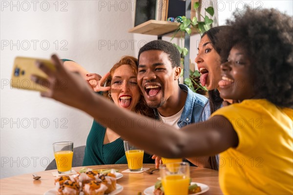 Multiethnic friends at a breakfast with orange juice and muffins at home, selfie smiling and having fun