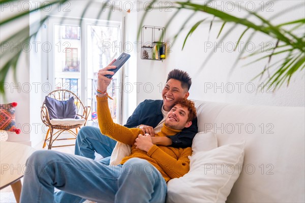 Beautiful gay couple being romantic indoors at home on the sofa, taking a selfie, lgbt concept