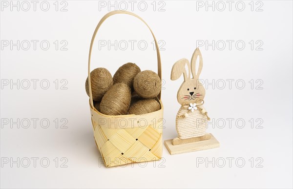 Easter composition. Basket with eggs wrapped in jute twine. Wooden bunny on the side. White background. Studio