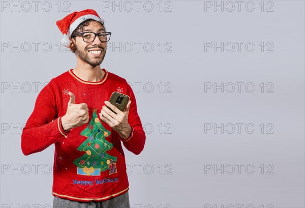 Smiling guy in christmas hat using cellphone with thumb up. Smiling young man in christmas hat using cellphone and giving thumbs up. People in santa hat using phone and doing ok gesture