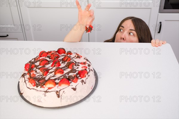 Young woman hiding behind the table eating strawberry shortcake in her home kitchen