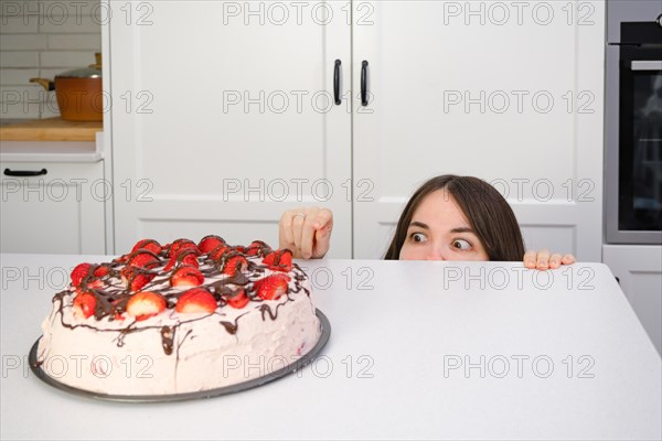 Young woman hiding behind the table eating strawberry shortcake in her home kitchen