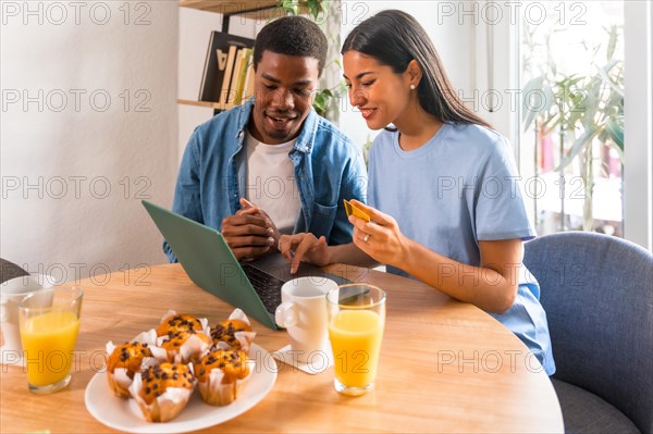 Multi-ethnic couple making purchase online with the computer while having breakfast, next to the window