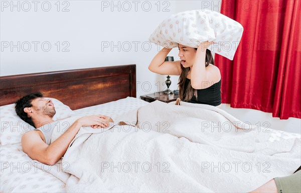 Suffering wife covering her ears while her husband snores. Angry woman holding pillow and snoring husband, Snoring man in bedroom and wife covers her ears with pillow