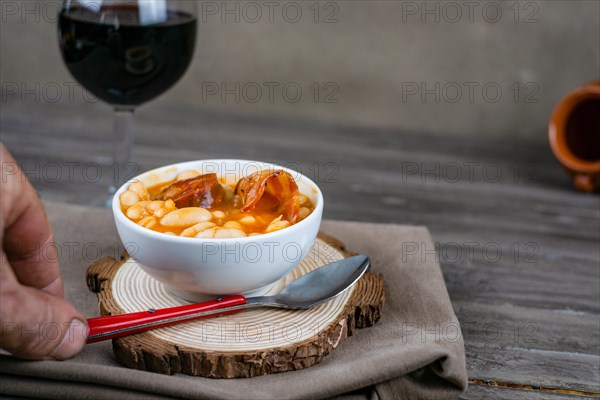 White beans stewed with chorizo in white ceramic bowl on wooden table with a glass of red wine