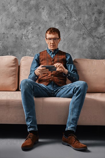 Serious middle aged man sitting on sofa and watching video on smartphone