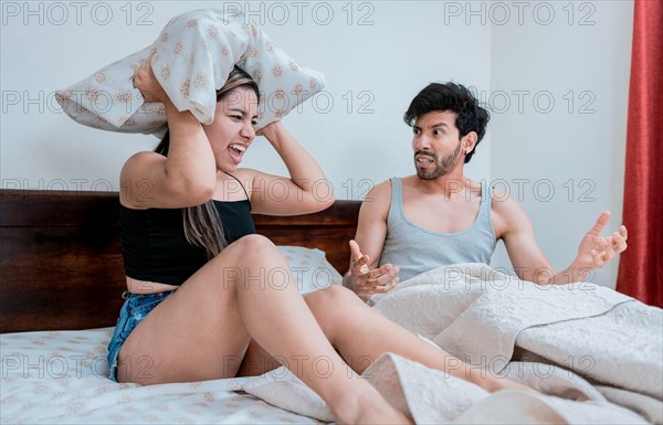 Young couple arguing in bed with the woman screaming and the man covering his ears with a pillow. Upset woman yelling at husband in bedroom bed