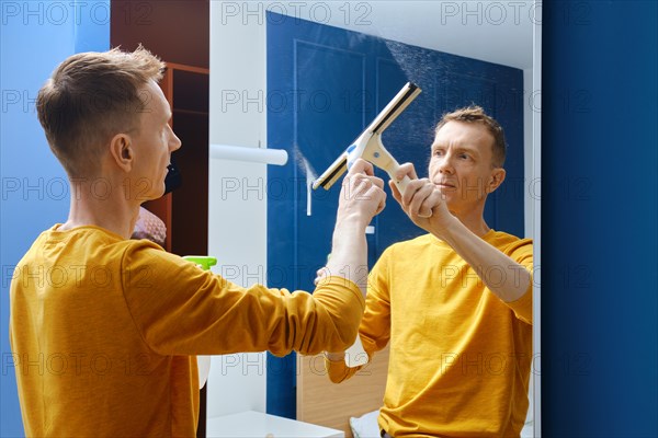 Middle-aged man cleaning mirror at home with rubber mop