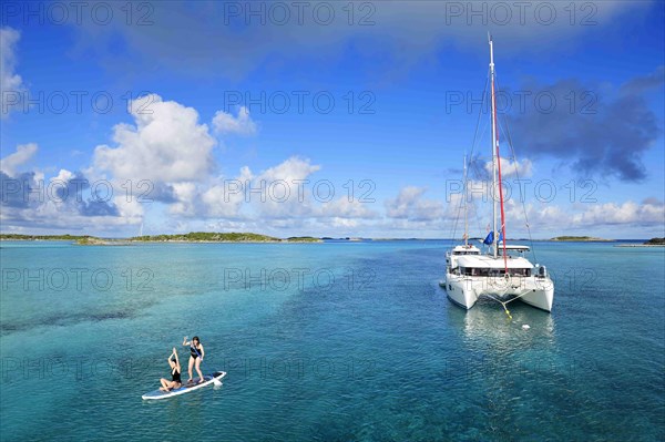 Women paddle SUP and do yoga in front of a sailing catamaran in the harbour of Warderick Wells