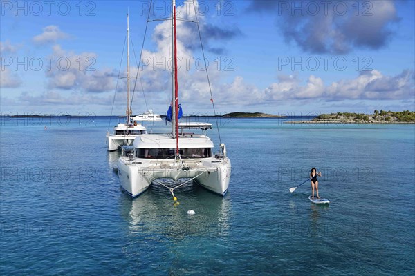 A woman paddles a SUP in front of a sailing catamaran in the harbour of Warderick Wells