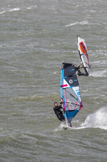 Two recreational windsurfers in black wetsuits practising classic windsurfing along the North Sea coast in windy weather during winter storm