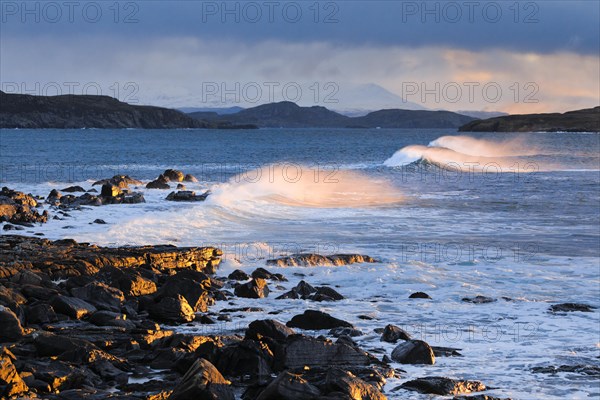 Large waves crash in a winter storm and the swirling spray is illuminated by the warm light of the evening sun